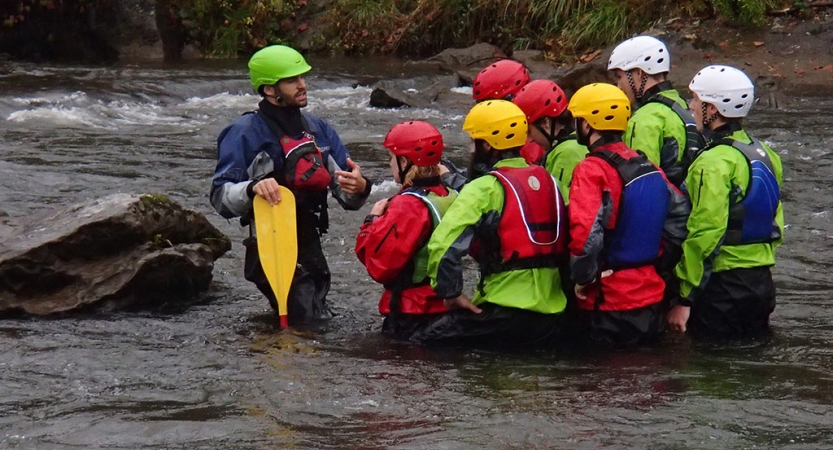 A group of people wearing safety gear stand in waist deep water, listening to an instructor speak.
